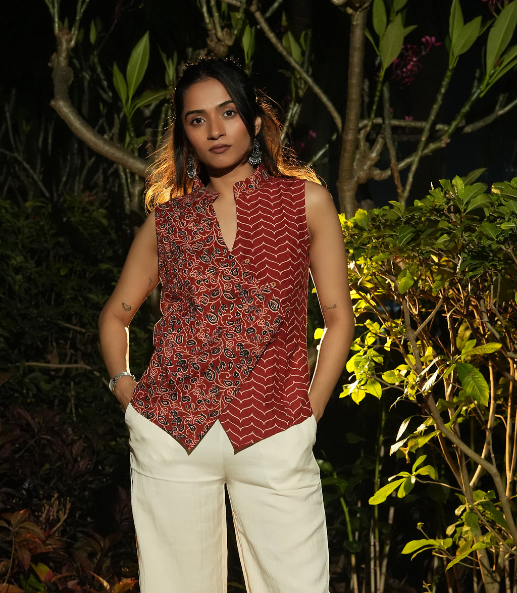 Woman wearing a patterned Maroon overlap top and white pants standing among green foliage.