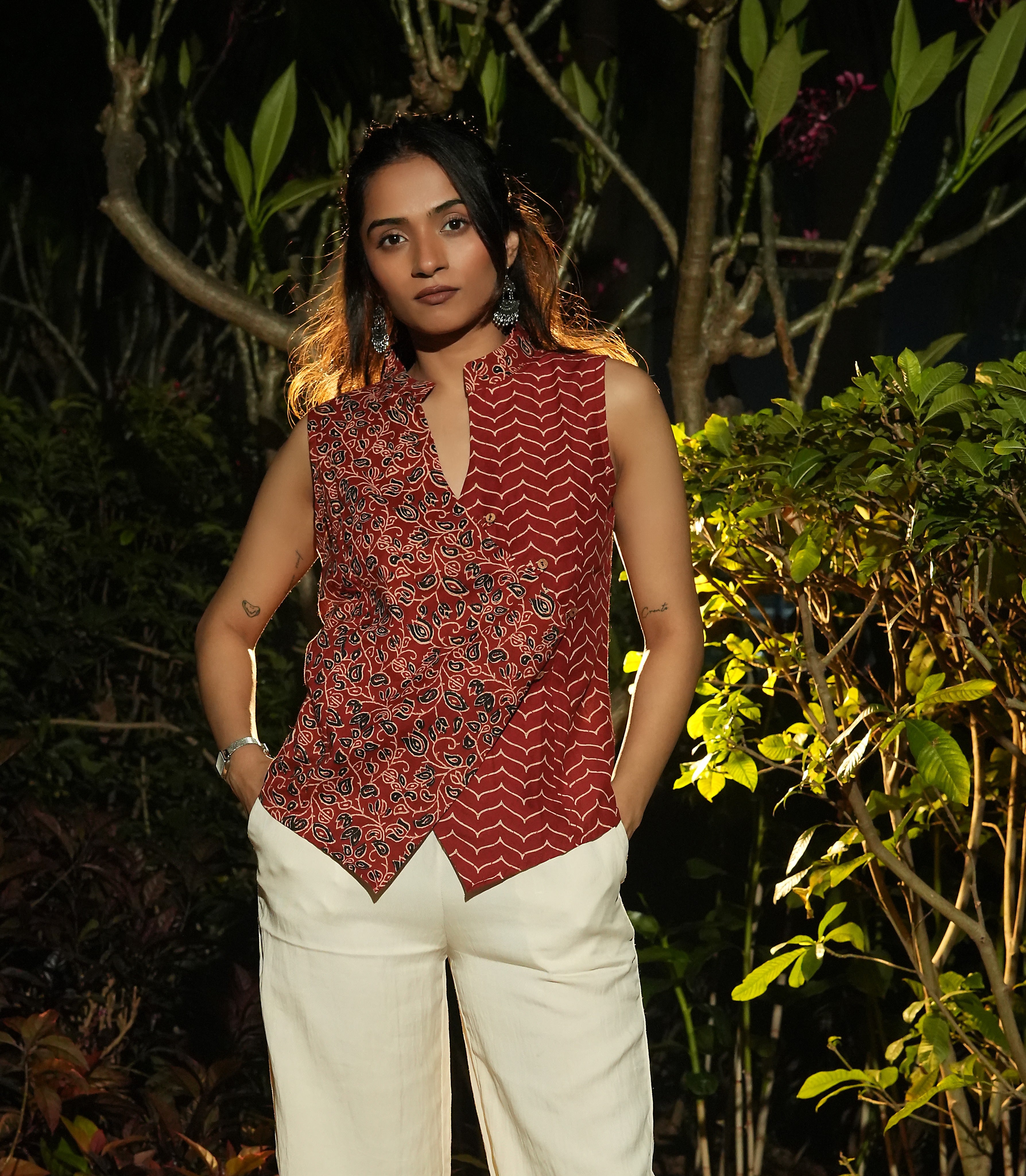 Woman wearing a patterned Maroon overlap top and white pants standing among green foliage.